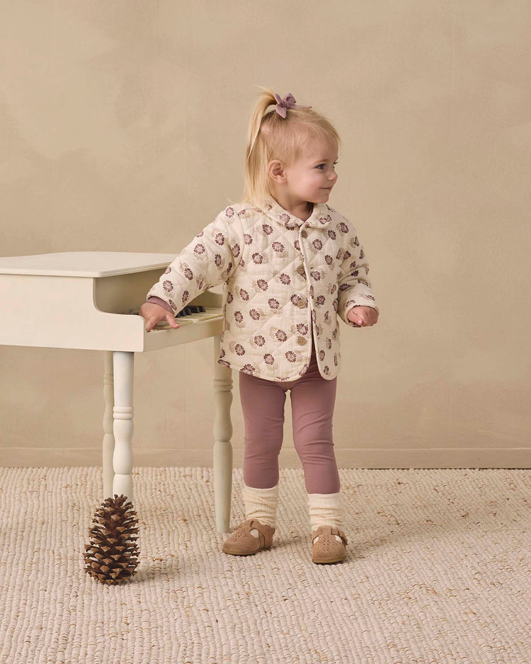 A young child with light hair tied with a bow stands by a small white table, wearing the quincy mae quilted jacket in fall floral and pink leggings. A large pinecone rests on the floor nearby.
