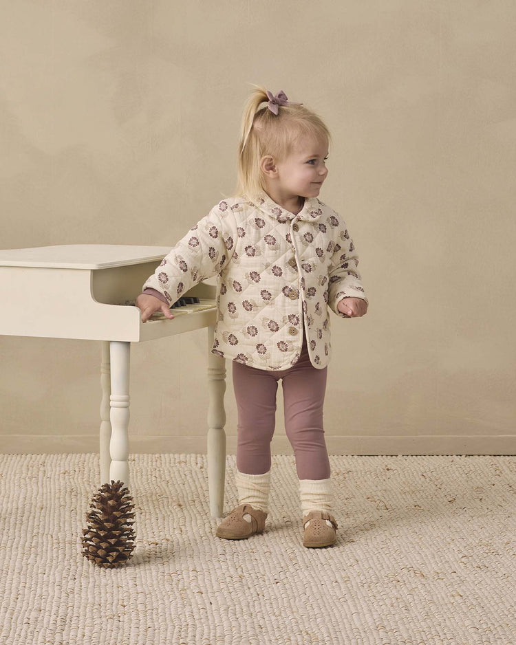 A young child with light hair tied with a bow stands by a small white table, wearing the quincy mae quilted jacket in fall floral and pink leggings. A large pinecone rests on the floor nearby.