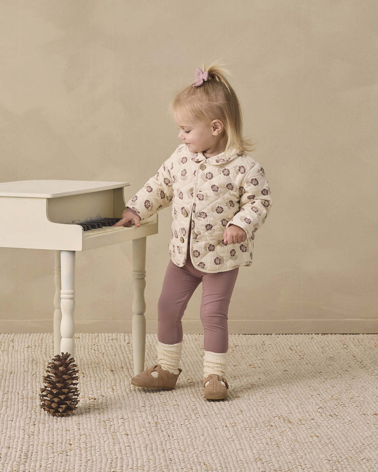 A young child wears the quincy mae quilted jacket in fall floral and mauve leggings, standing beside a small white piano as she touches its keys. A pinecone sits on the textured rug near her feet, all set against a soft, neutral background.