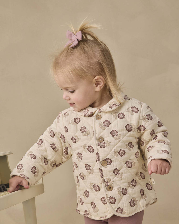 A toddler with a pink bow in her blonde hair wears the quincy mae quilted jacket in fall floral, standing beside a small white piano and looking down.