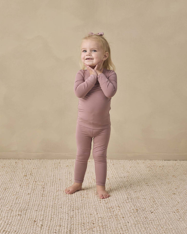 A young child with blonde hair, wearing the quincy mae bamboo pajama set in mulberry—which includes a slim-fit long sleeve tee and matching leggings—stands barefoot on a textured cream rug, smiling with hands on their cheeks against a beige background.