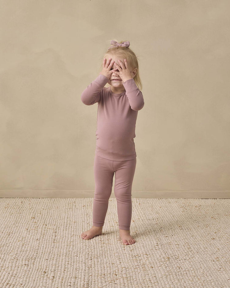 A young child in a quincy mae bamboo pajama set in mulberry with a bow stands barefoot on a textured rug, covering their eyes with both hands, against a plain beige background.