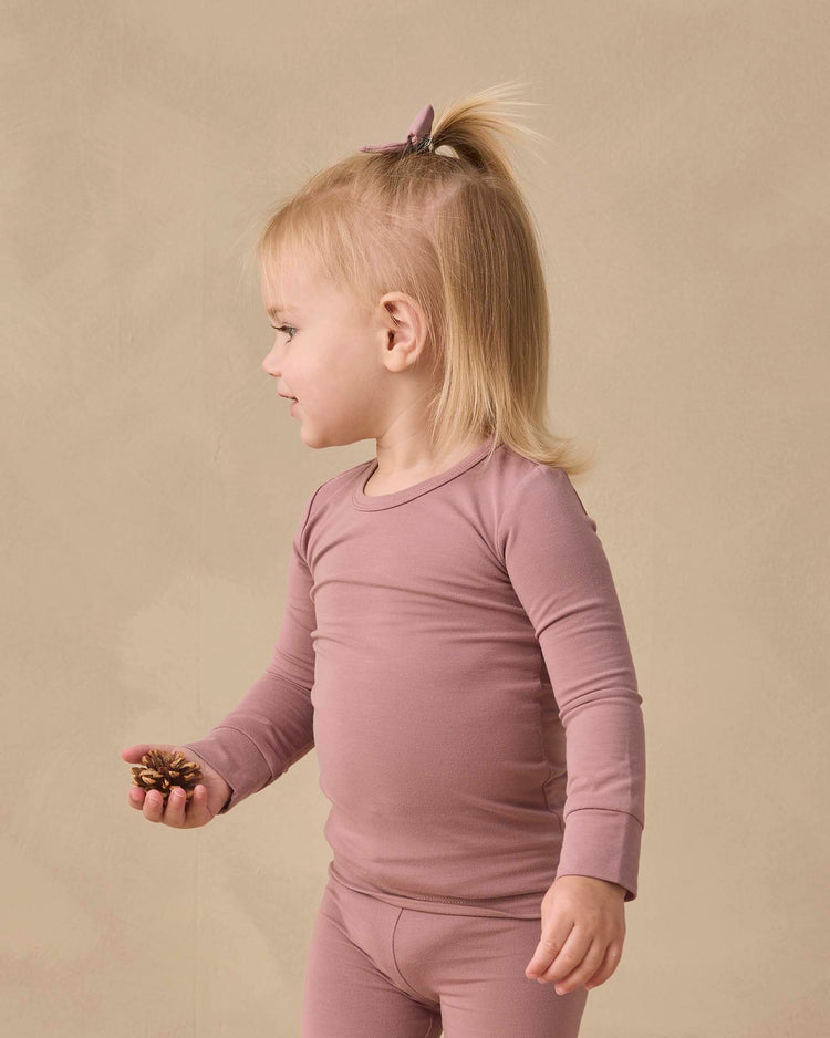 A toddler with blond hair in a ponytail wears the quincy mae bamboo pajama set in mulberry, standing sideways and holding a pine cone against a beige background.