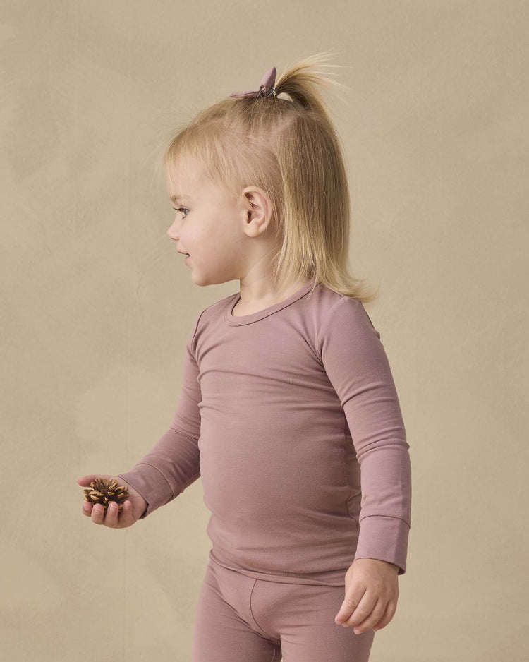 A toddler with blond hair in a ponytail wears the quincy mae bamboo pajama set in mulberry, standing sideways and holding a pine cone against a beige background.