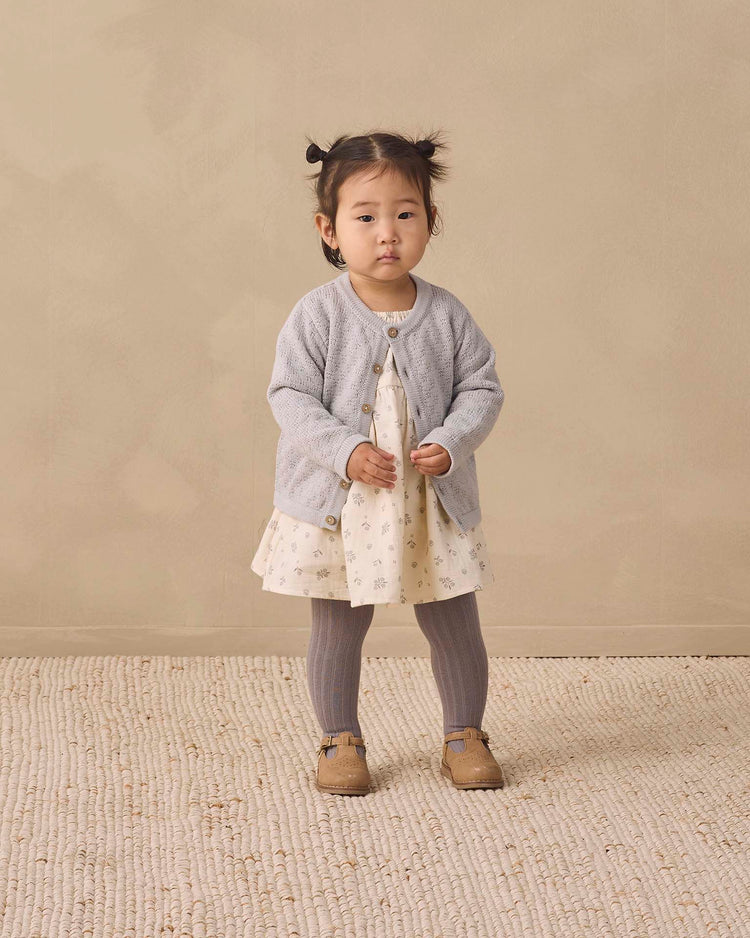 A toddler with pigtails stands on a textured carpet against a beige background, wearing a light-colored dress with a gray cardigan, Quincy Mae tights in slate (organic cotton), and brown shoes.
