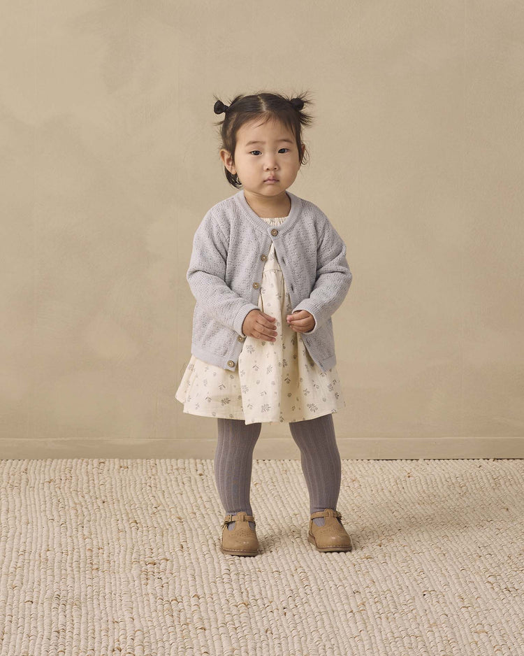 A toddler with pigtails stands on a textured carpet against a beige background, wearing a light-colored dress with a gray cardigan, Quincy Mae tights in slate (organic cotton), and brown shoes.