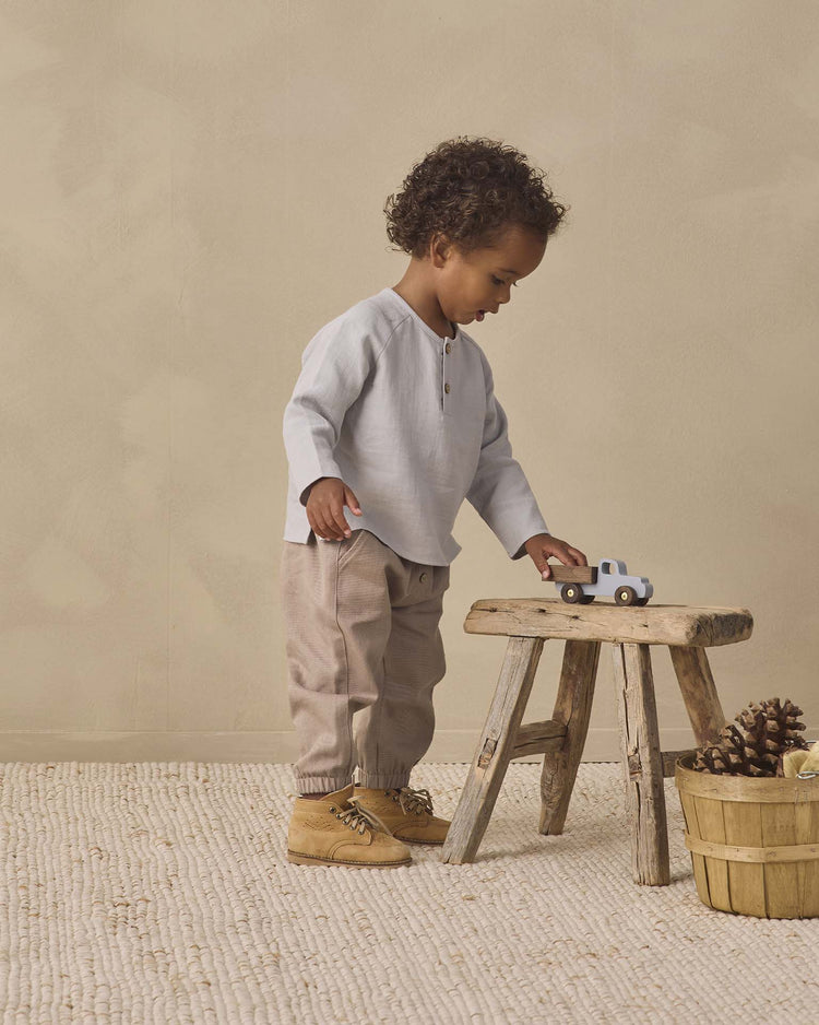 A toddler stands on a cream rug, playing with a toy truck atop a wooden stool. Nearby, a basket of pinecones rests on the floor. The child wears beige pants, tan shoes, and the quincy mae zion shirt in cloud—an organic cotton top in a relaxed fit—set against a neutral wall.