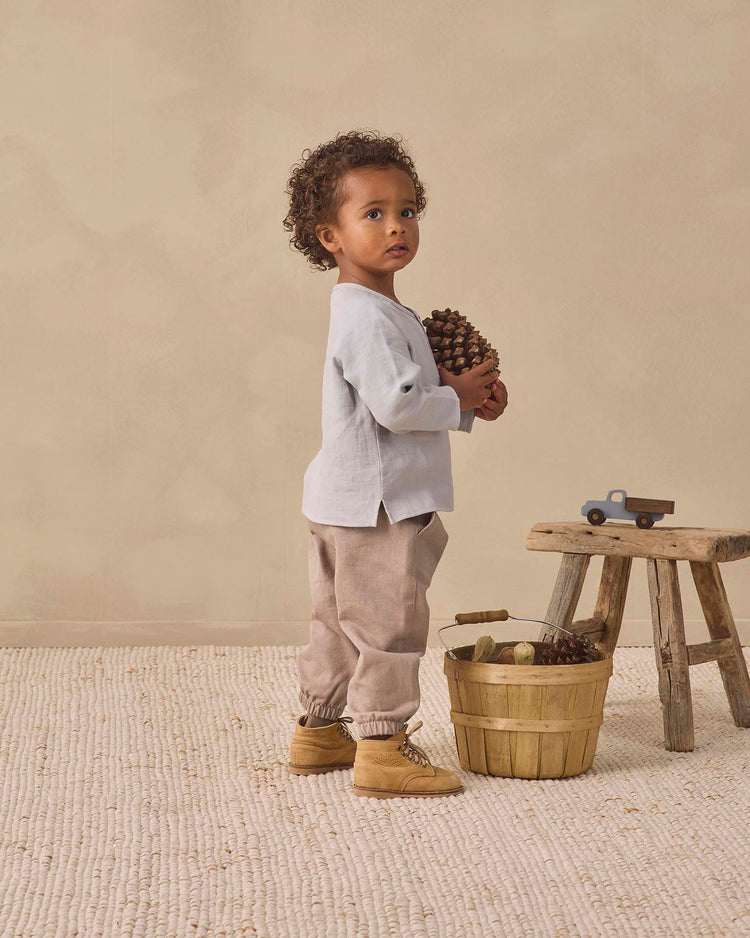 A young child stands on a light rug holding a large pinecone, dressed in the quincy mae zion shirt in cloud, which adds to the cozy feel of the scene. A wooden stool with a toy car and a basket filled with more pinecones and wood are nearby, set against a beige background.