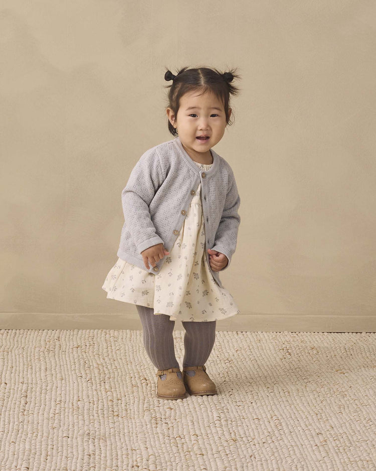 A young child with pigtails stands on a textured rug, wearing a light cardigan, a cream patterned dress, quincy mae tights in slate made from organic cotton, and brown shoes, against a plain beige background.