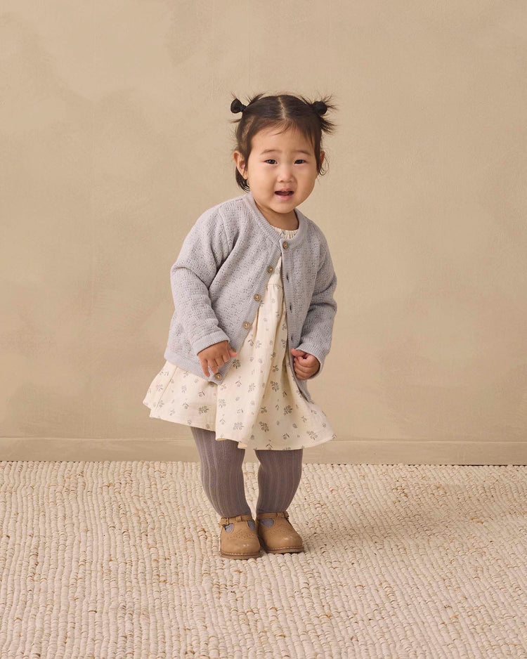 A young child with pigtails stands on a textured beige rug, wearing the Quincy Mae knit cardigan in cloud—crafted from organic cotton with coconut buttons—paired with a patterned dress, gray tights, and brown shoes, set against a neutral beige background.