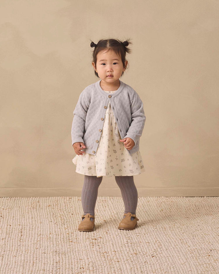 A young child stands on a textured rug wearing a cream dress with a floral pattern, a light gray cardigan, quincy mae tights in slate, and brown shoes, against a neutral beige background.