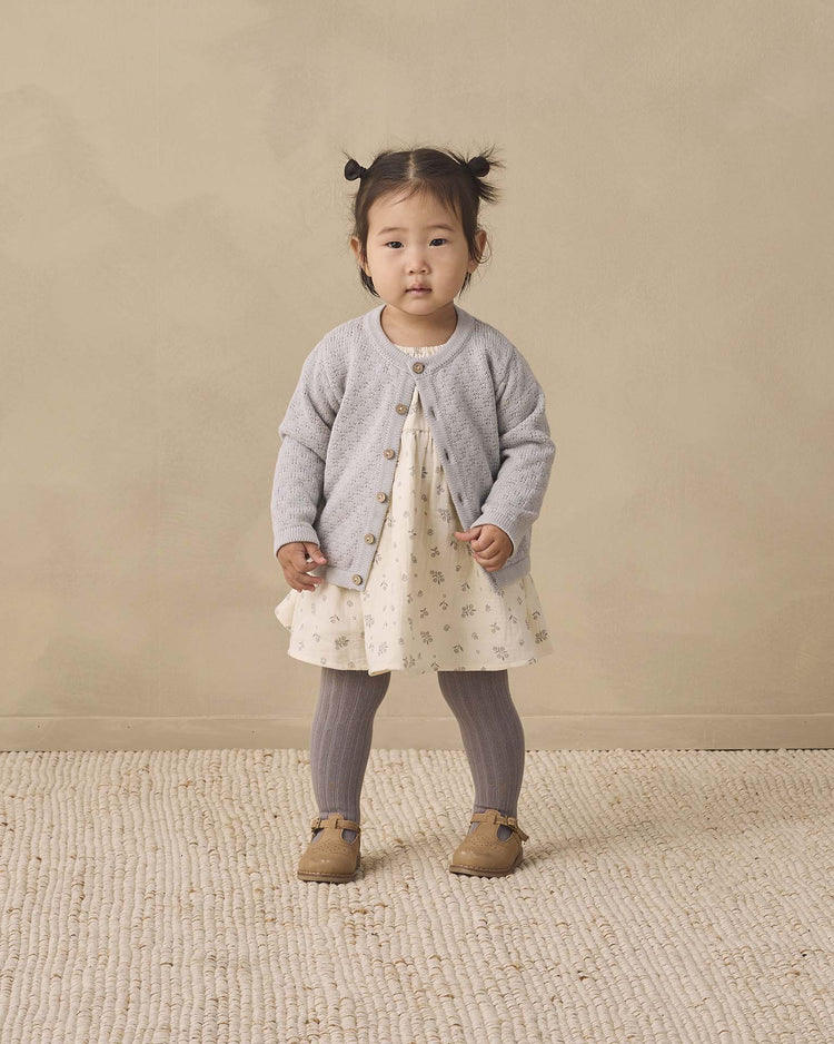 A young child stands on a textured rug wearing a cream dress with a floral pattern, a light gray cardigan, quincy mae tights in slate, and brown shoes, against a neutral beige background.