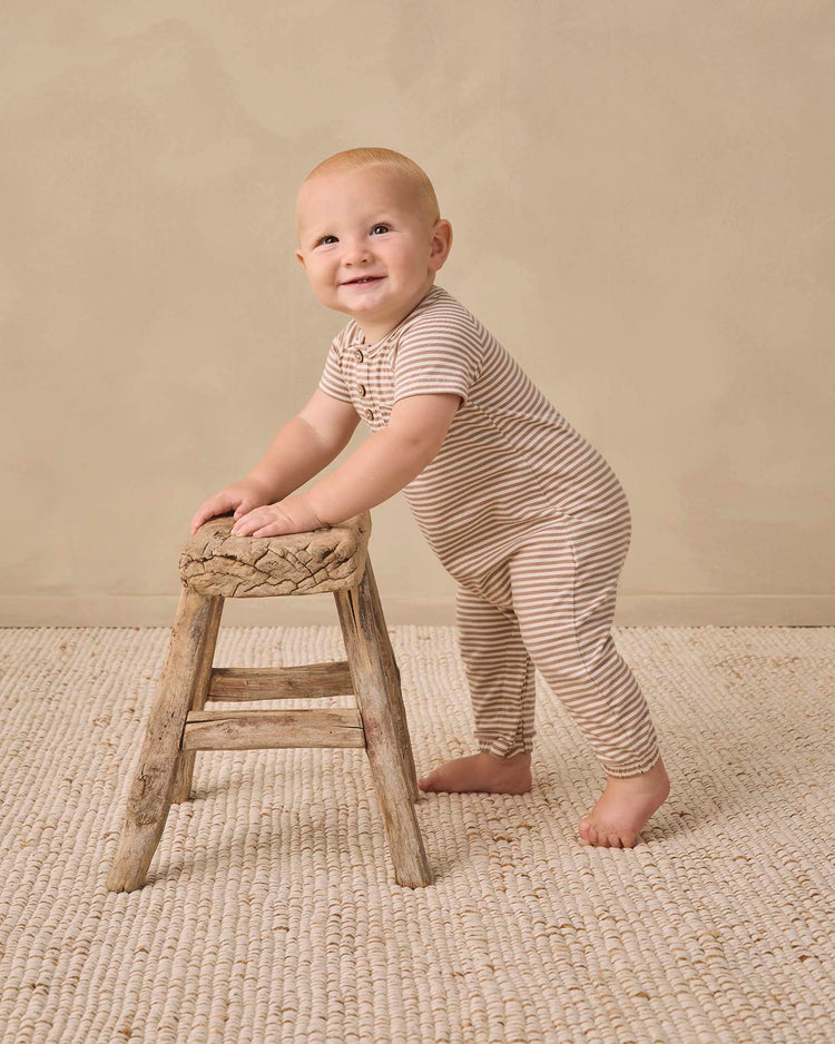 A smiling baby wearing the quincy mae short sleeve jumpsuit in almond stripe stands while holding onto a small, rustic wooden stool on a textured cream carpet with a plain beige background.