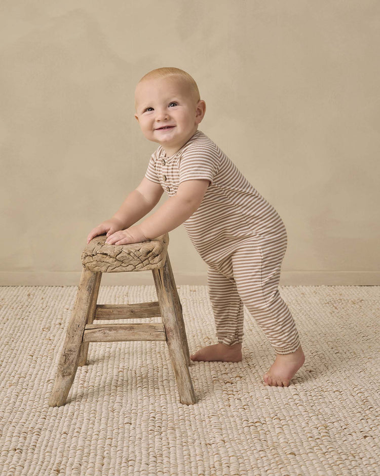 A smiling baby wearing the quincy mae short sleeve jumpsuit in almond stripe stands while holding onto a small, rustic wooden stool on a textured cream carpet with a plain beige background.