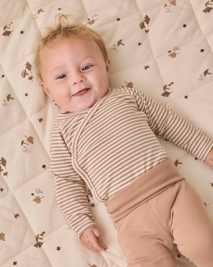A smiling baby with light hair wears the Quincy Mae side snap bodysuit + pant set in almond stripe—featuring a beige striped top and tan footed pants—while lying on a cream quilted blanket decorated with brown animal prints.