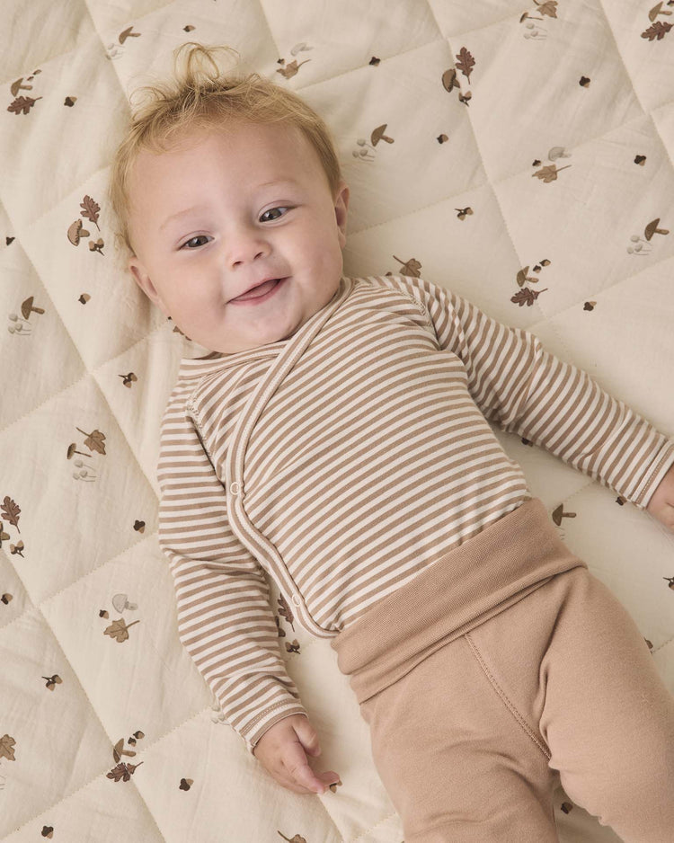 A smiling baby with light hair wears the Quincy Mae side snap bodysuit + pant set in almond stripe—featuring a beige striped top and tan footed pants—while lying on a cream quilted blanket decorated with brown animal prints.