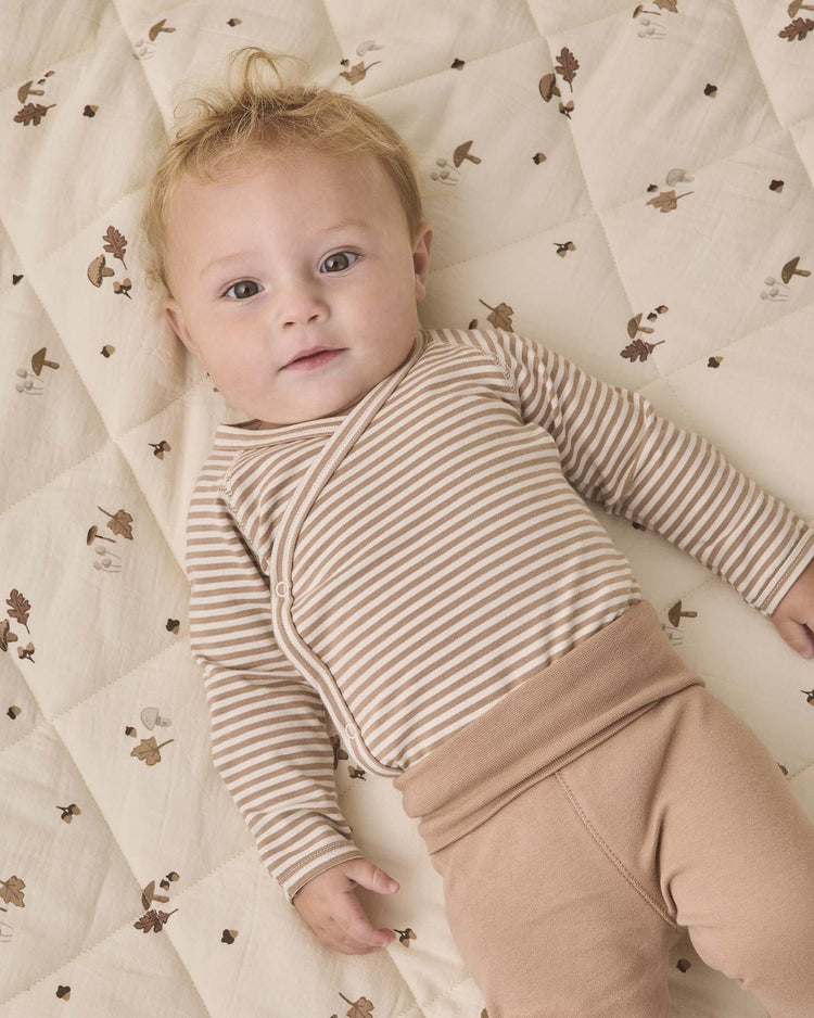 A baby with light hair lies on a quilted blanket adorned with a whimsical pattern of small animals, acorns, and leaves, wearing the quincy mae side snap bodysuit + pant set in almond stripe—an organic cotton ensemble featuring beige and white stripes paired with cozy tan footed pants.