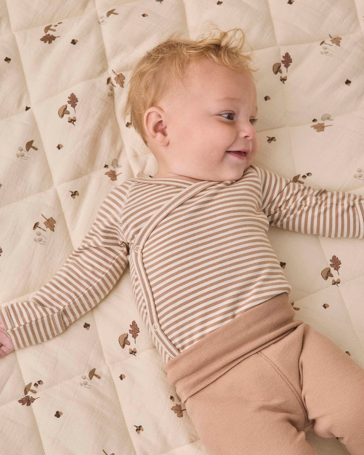 A smiling blonde baby lies on a beige quilt with brown leaf patterns, wearing the quincy mae side snap bodysuit + pant set in almond stripe—featuring a cozy organic cotton striped top and matching tan footed pants.