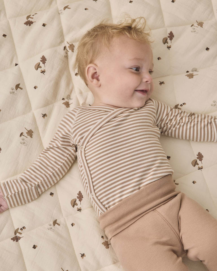 A smiling blonde baby lies on a beige quilt with brown leaf patterns, wearing the quincy mae side snap bodysuit + pant set in almond stripe—featuring a cozy organic cotton striped top and matching tan footed pants.