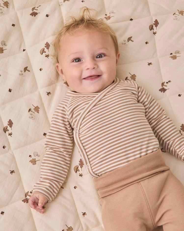 A smiling baby with blond hair wears the Quincy Mae side snap bodysuit + pant set in almond stripe—featuring a beige and white striped bodysuit and tan footed pants—while lying on a cream quilt with a nature-themed pattern.