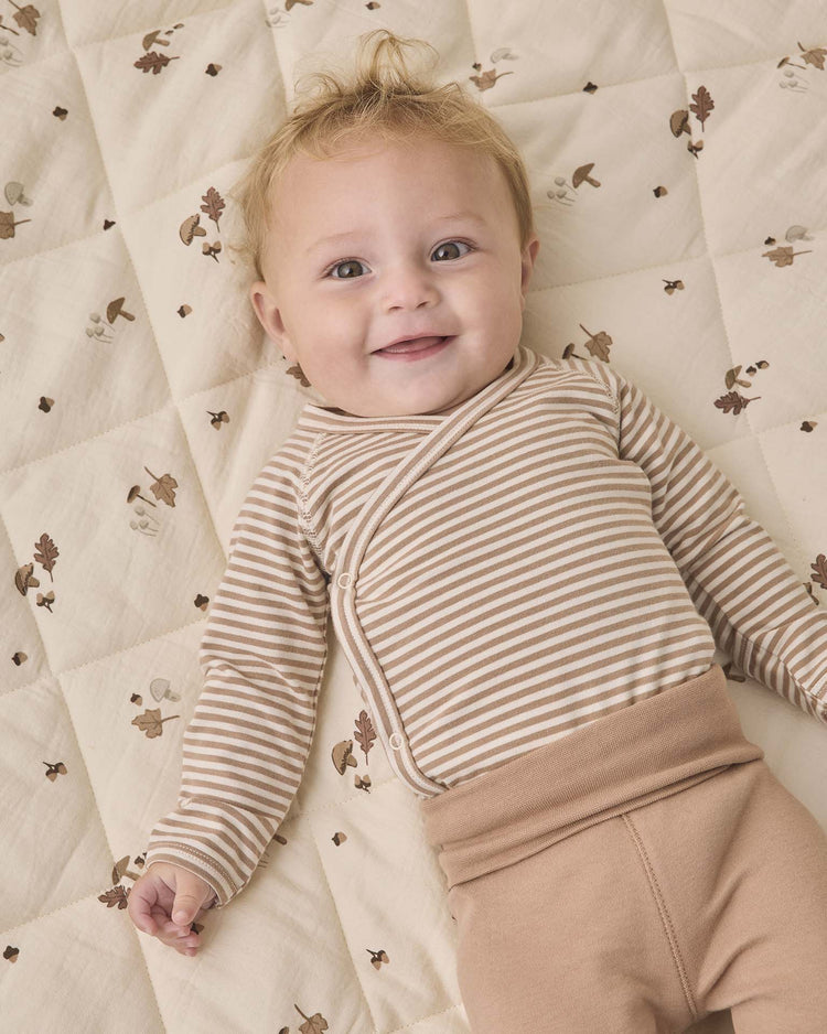 A smiling baby with blond hair wears the Quincy Mae side snap bodysuit + pant set in almond stripe—featuring a beige and white striped bodysuit and tan footed pants—while lying on a cream quilt with a nature-themed pattern.