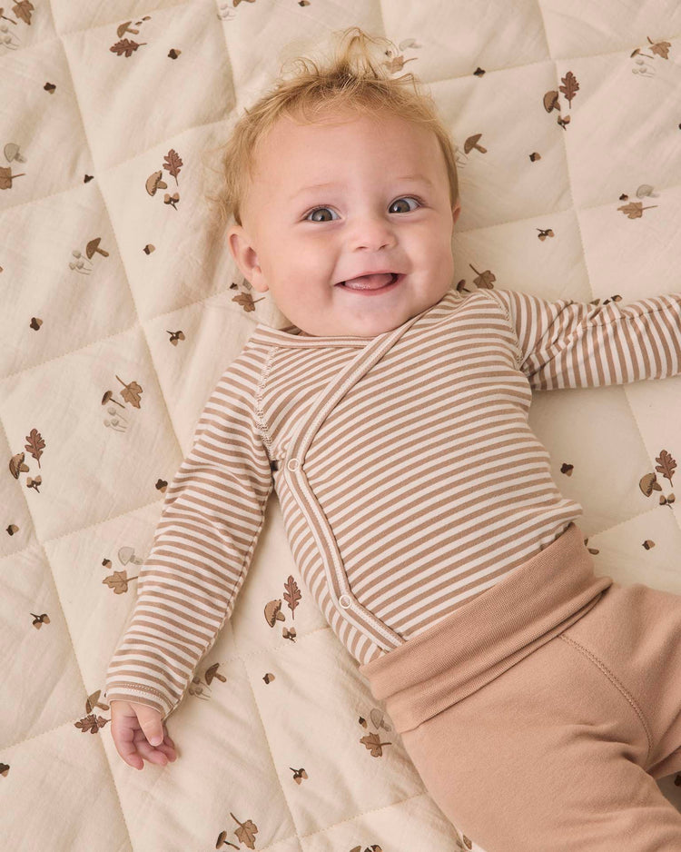 A smiling baby with curly hair wears the Quincy Mae side snap bodysuit + pant set in almond stripe, lying on a quilted blanket patterned with acorns and oak leaves.