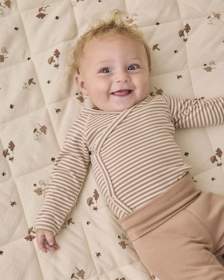 A smiling baby with curly hair wears the Quincy Mae side snap bodysuit + pant set in almond stripe, lying on a quilted blanket patterned with acorns and oak leaves.