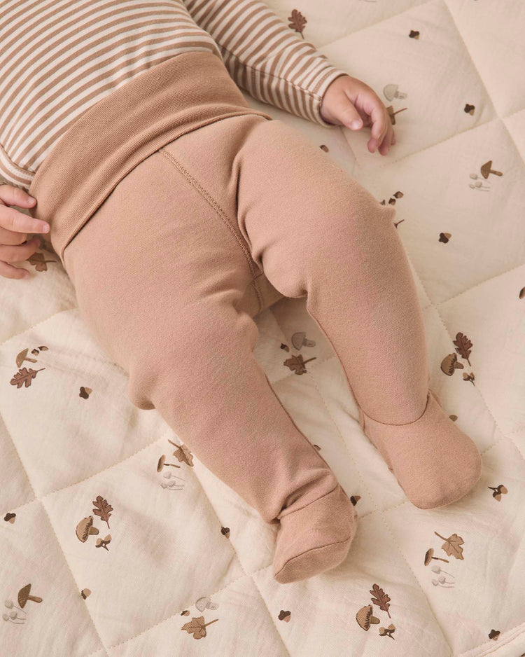 A baby wearing the quincy mae side snap bodysuit + pant set in almond stripe lies on a cream quilt decorated with small mushrooms and leaves. Only the baby’s torso, arms, and legs are visible.