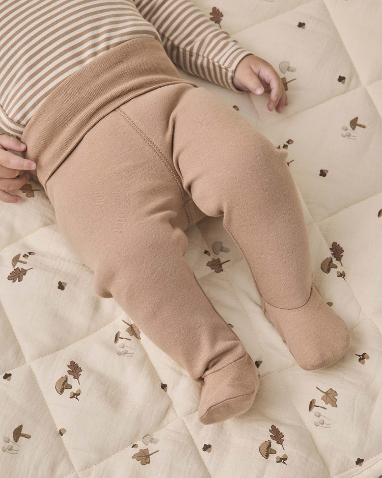 A baby wearing the quincy mae side snap bodysuit + pant set in almond stripe lies on a cream quilt decorated with small mushrooms and leaves. Only the baby’s torso, arms, and legs are visible.