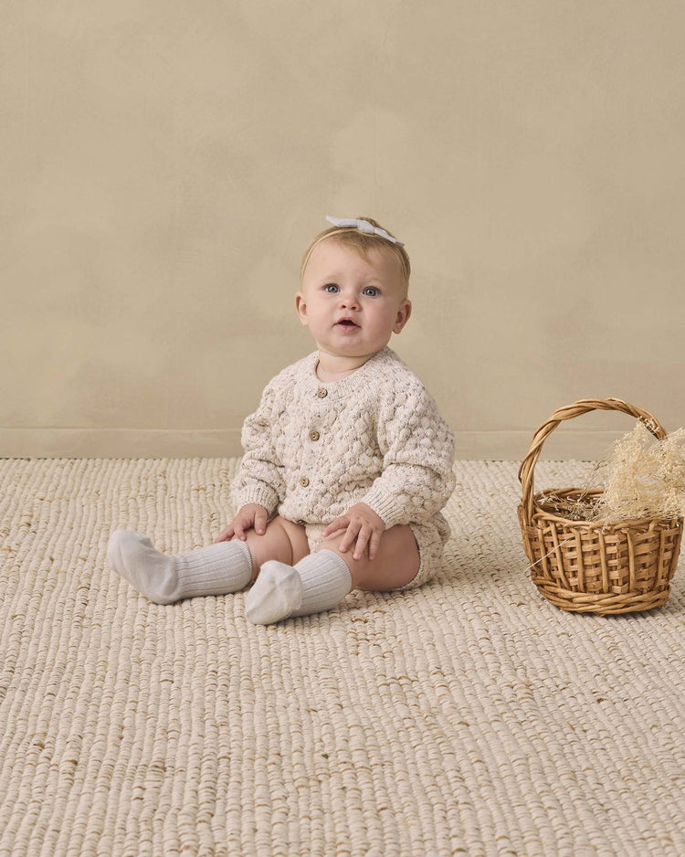 A baby wearing the quincy mae bubble knit cardigan in natural confetti sits on a textured beige rug next to a wicker basket with straw, against a neutral background.