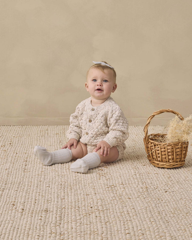 A baby wearing the quincy mae bubble knit cardigan in natural confetti, paired with white socks and a bow, sits on a textured rug next to a wicker basket filled with dried grass, set against a beige background.