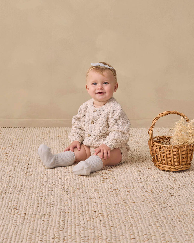 A baby wearing the quincy mae bubble knit cardigan in natural confetti, cozy socks, and a white bow sits on a textured beige rug next to a small wicker basket filled with straw, against a plain beige background. Made from organic cotton, the sweater provides an extra layer of softness and comfort to this charming scene.