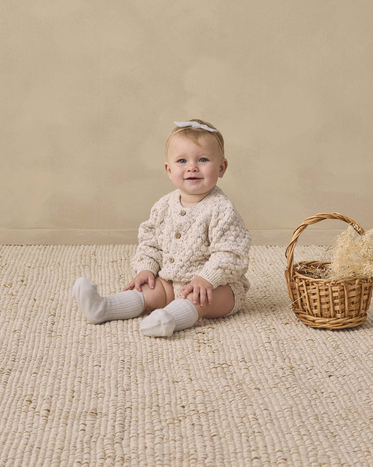 A baby wearing the quincy mae bubble knit cardigan in natural confetti, cozy socks, and a white bow sits on a textured beige rug next to a small wicker basket filled with straw, against a plain beige background. Made from organic cotton, the sweater provides an extra layer of softness and comfort to this charming scene.