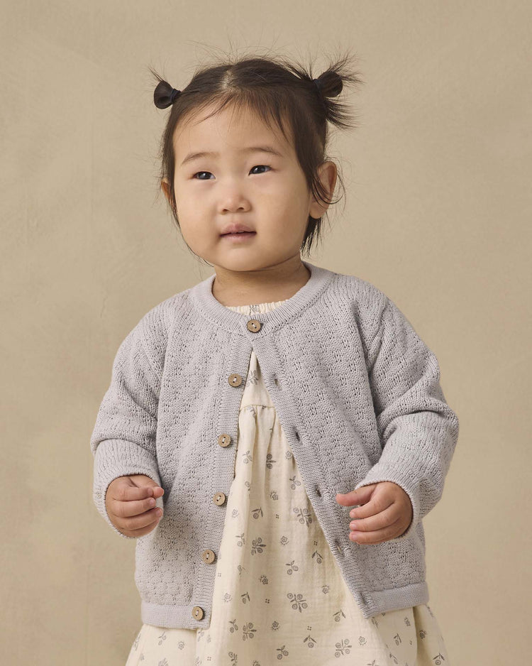 A young child with pigtails wears the quincy mae knit cardigan in cloud, featuring coconut buttons, over a cream dress patterned with small flowers while standing against a plain beige background.