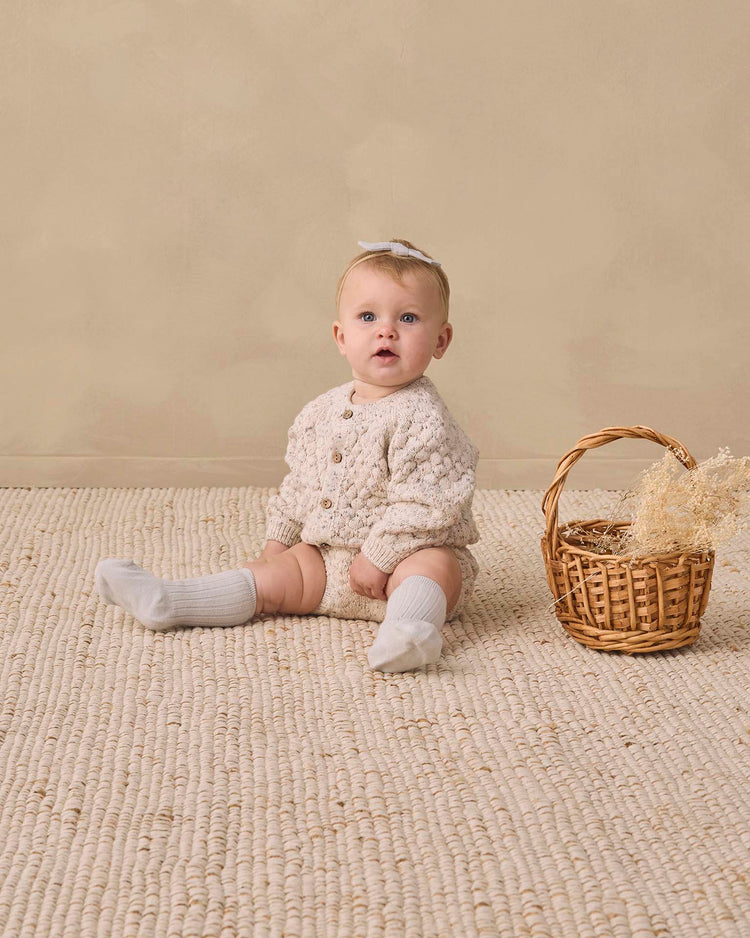 A baby in a quincy mae bubble knit cardigan in natural confetti and white socks sits on a textured rug next to a small wicker basket with dried flowers, against a beige background.