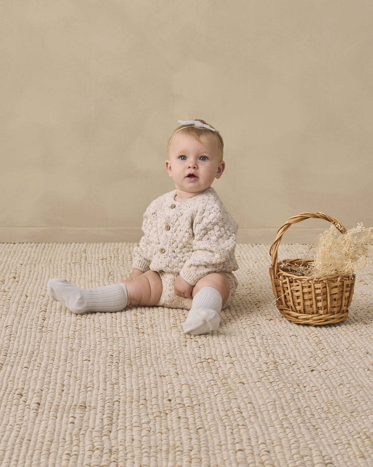 A baby in a quincy mae bubble knit cardigan in natural confetti and white socks sits on a textured rug next to a small wicker basket with dried flowers, against a beige background.
