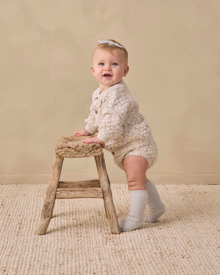 A baby dressed in a cream knit outfit and headband stands holding a rustic wooden stool on a textured beige rug, with a neutral background. Their look is completed with the cozy quincy mae socks set in slate + cloud, adding an extra touch of warmth and charm to the adorable ensemble.