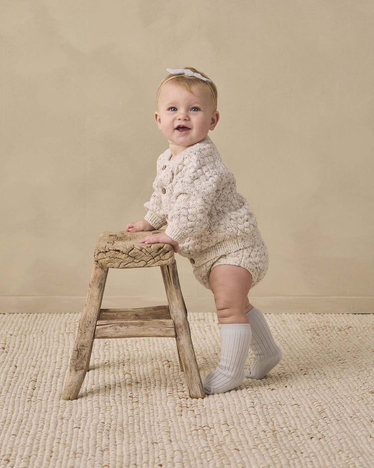 A baby wearing quincy mae's knit bloomer in natural confetti, paired with a cream knitted outfit, headband, and knee-high socks, stands holding a rustic wooden stool on a textured rug against a beige wall.