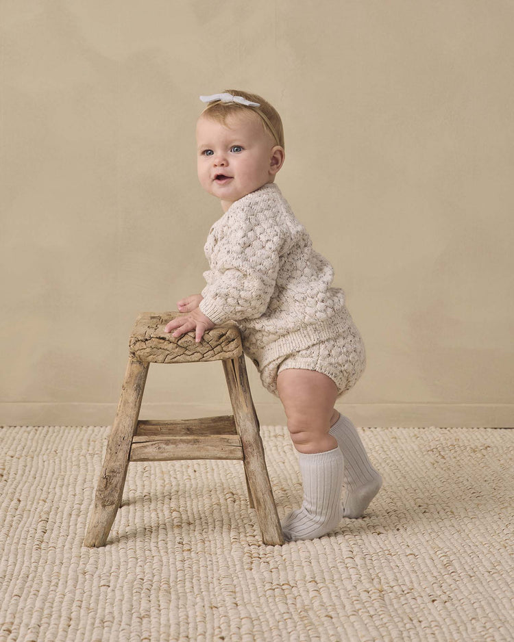 A baby wearing the quincy mae knit bloomer in natural confetti with a cream knit outfit and white knee-high socks stands while holding onto a rustic wooden stool, on a textured beige rug against a neutral wall.