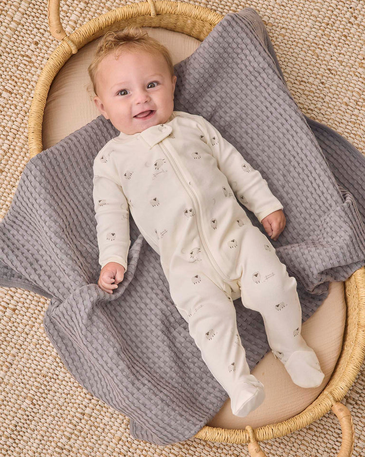 A smiling baby wearing the quincy mae zip long sleeve sleeper footie in sheep lies on a gray textured blanket in a round woven basket on a beige woven rug.