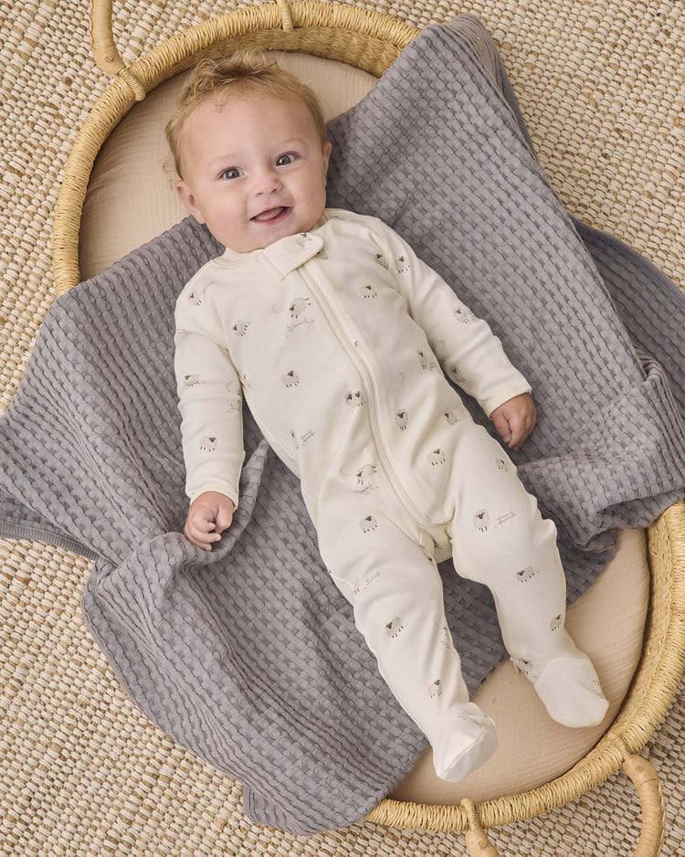 A smiling baby wearing the quincy mae zip long sleeve sleeper footie in sheep lies on a gray textured blanket in a round woven basket on a beige woven rug.