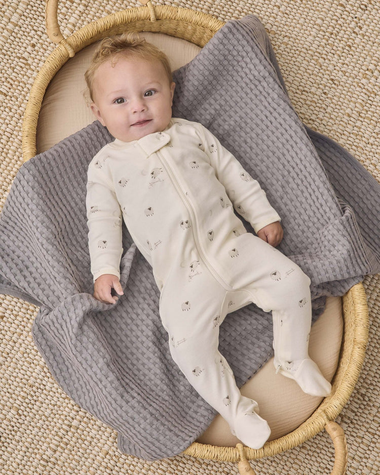 A baby wearing the quincy mae zip long sleeve sleeper footie in sheep rests on a gray textured blanket in a woven basket, gazing upward with a gentle smile.