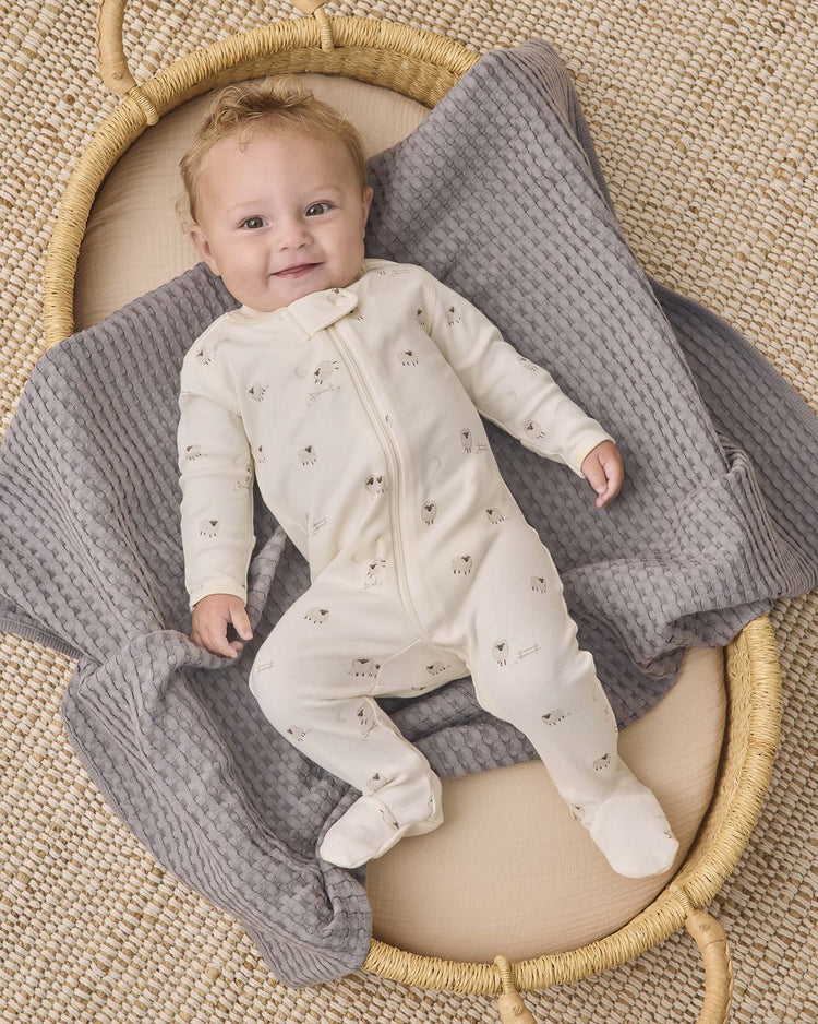 A smiling baby lies on a gray textured blanket in a woven basket, wearing the Quincy Mae zip long sleeve sleeper footie in sheep—a perfect choice for comfortable baby sleepwear.