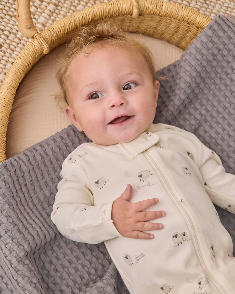 A baby with light brown hair, wearing the quincy mae zip long sleeve sleeper footie in sheep—a cream-colored organic cotton sleeper decorated with small sheep prints—lies on a textured gray blanket inside a wicker basket, looking up and smiling.