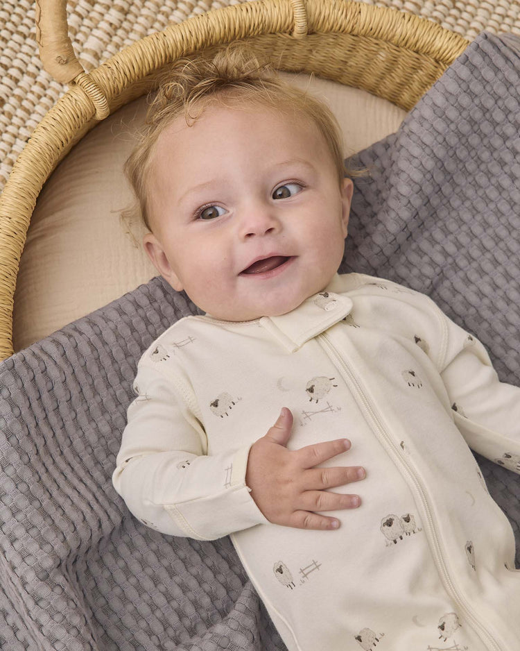 A baby with light brown hair, wearing the quincy mae zip long sleeve sleeper footie in sheep—a cream-colored organic cotton sleeper decorated with small sheep prints—lies on a textured gray blanket inside a wicker basket, looking up and smiling.