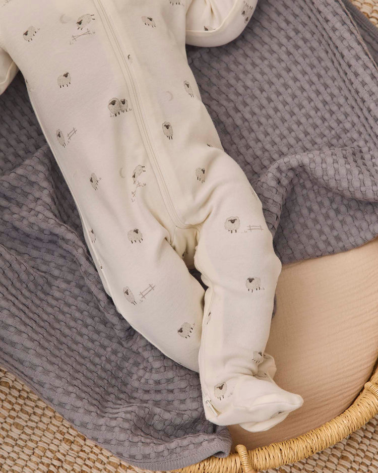 A baby dressed in the quincy mae zip long sleeve sleeper footie in sheep, made from white organic cotton with charming sheep prints, lies on a textured gray blanket in a woven basket. Only the lower half of the baby's body is visible, drawing attention to the cozy baby sleepwear.