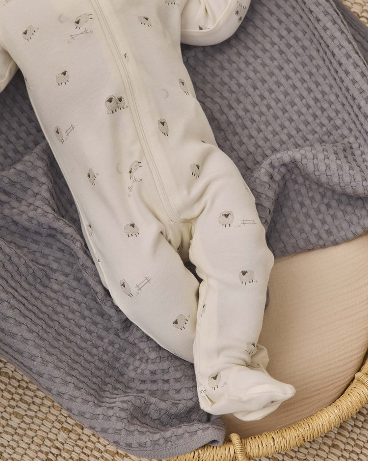A baby dressed in the quincy mae zip long sleeve sleeper footie in sheep, made from white organic cotton with charming sheep prints, lies on a textured gray blanket in a woven basket. Only the lower half of the baby's body is visible, drawing attention to the cozy baby sleepwear.
