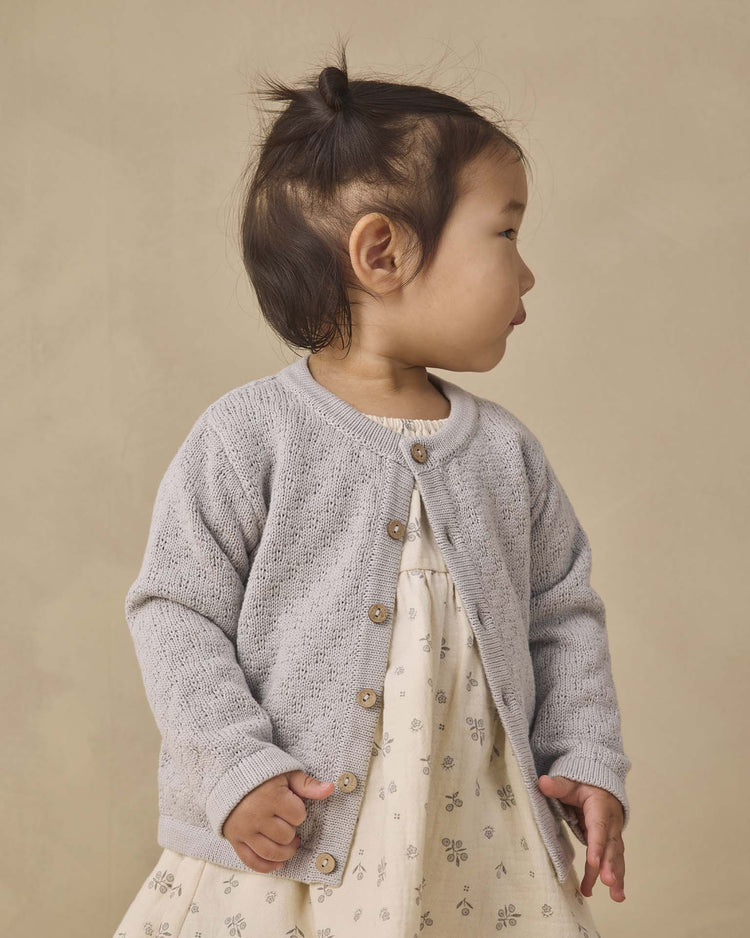 A young child with dark hair in a top knot wears the quincy mae knit cardigan in cloud, featuring coconut buttons, layered over a cream dress with a small floral pattern. She stands against a beige background, looking to the side.