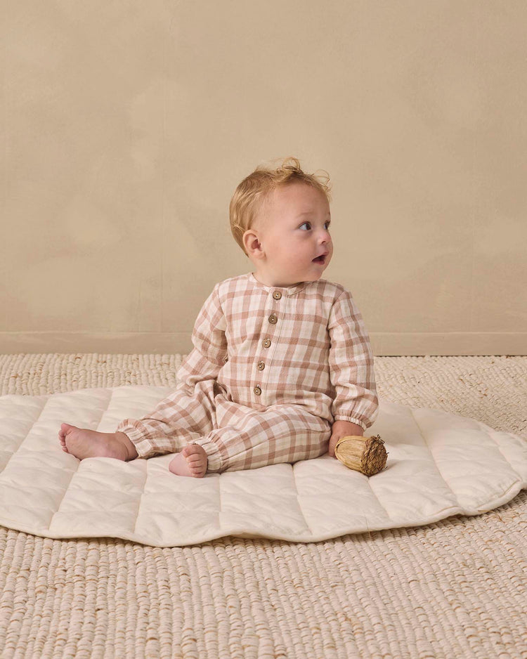 A baby wearing the quincy mae woven jumpsuit in rustic plaid sits on a quilted mat, holding a small toy against a neutral background.
