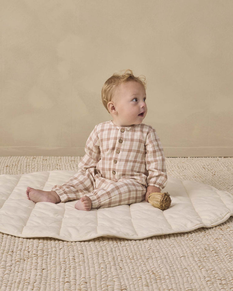 A baby wearing the quincy mae woven jumpsuit in rustic plaid sits on a quilted mat, holding a small toy against a neutral background.